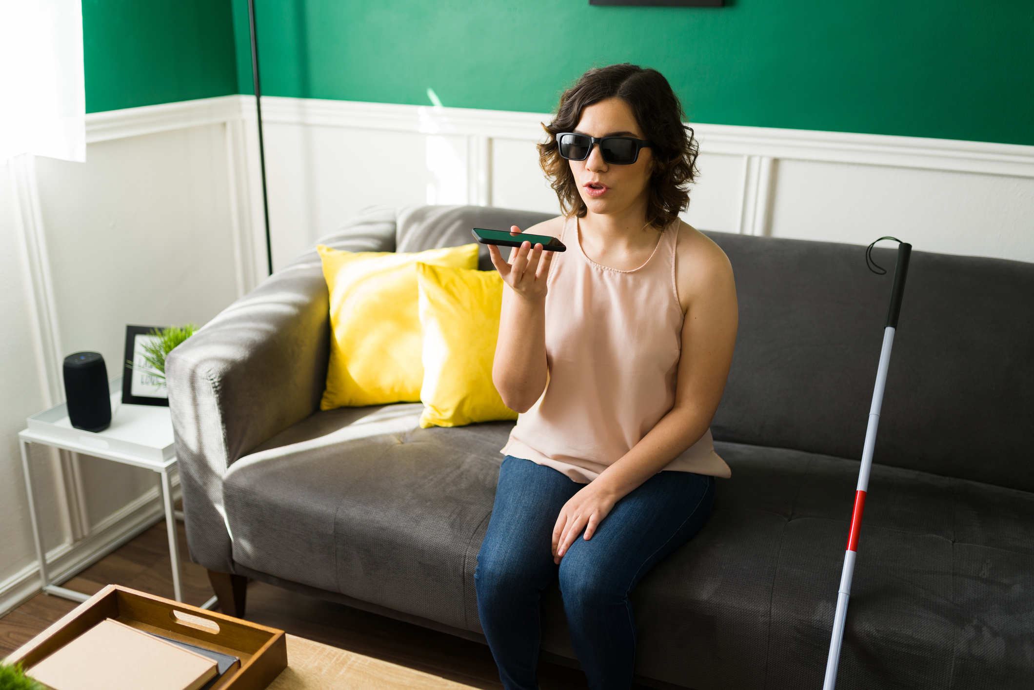 Foto de uma mulher cega sentada no sofá de uma sala com um celular na mão direta, próximo a sua boca. Ao seu lado está uma bengala.