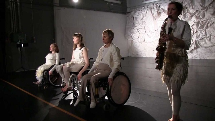 Em um palco, três cadeirantes em fila se apresentam ao lado de musicista com saxofone