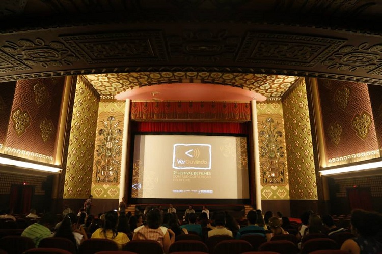 Plateia assiste a exibição de um filme em um teatro luxuoso. Na tela, está o nome do festival: Verouvindo