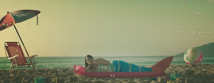 Cintia em uma praia, com guarda-sol e deitada na areia; ela usa calda de sereia e há bolhas de sabão em torno dela