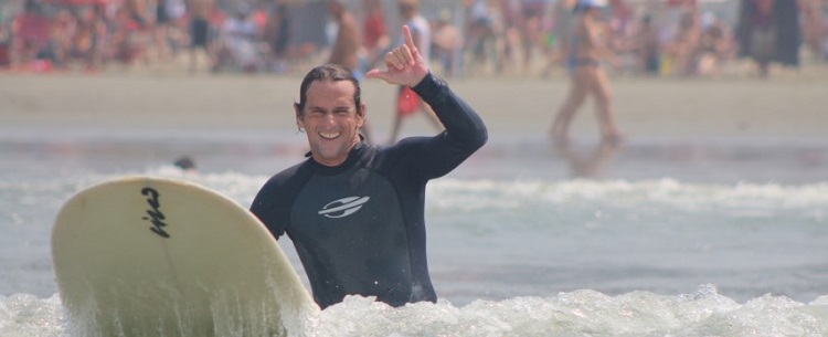 O surfista Cisco Araña em prancha, no mar, com a saudação havaiana de hang loose com as mãos (apenas polegar e mindinho levantados)
