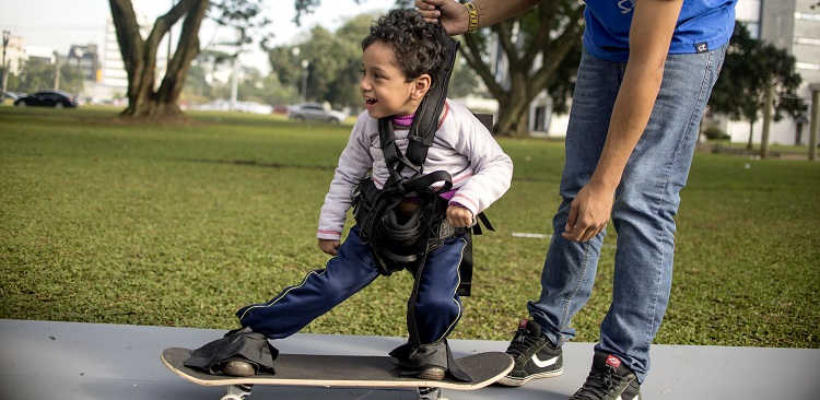 Menino de aproximadamente 6 anos, com deficiência motora, se equilibra em skate adaptado em um parque gramado e com árvores