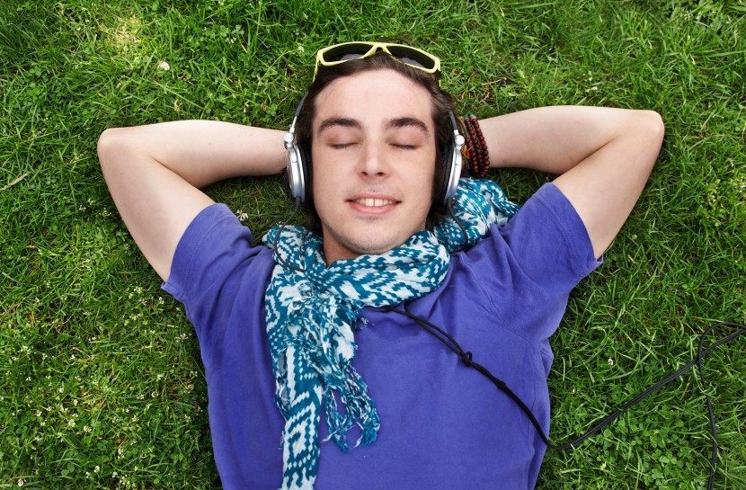 Rapaz está deitado de costas na grama, com as mãos atrás da cabeça. Com os olhos fechados, ele usa fones de ouvido e sorri.