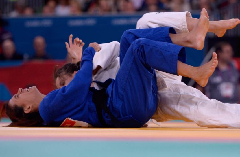 Duas judocas estão deitadas no tatame, lutando. Uma usa kimono azul, e a outra usa kimono branco.