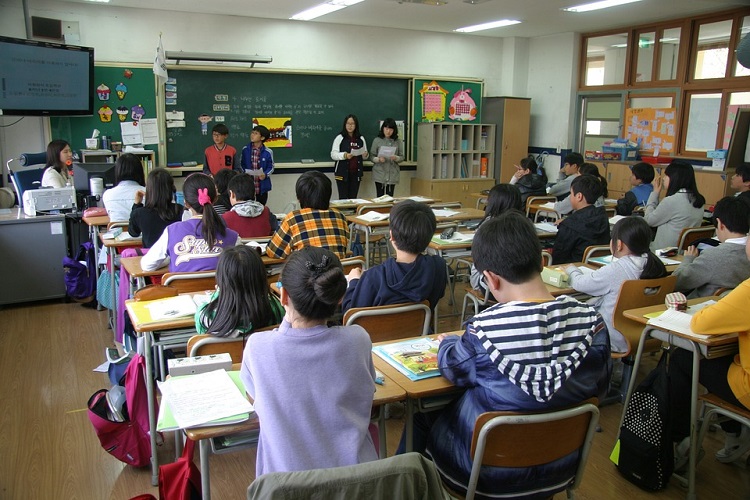Sala de aula vista do fundo, com uma professora lecionando e os alunos virados na direção dela