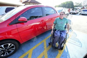 Em um estacionamento, um cadeirante de idade sorri ao lado de um veículo vermelho