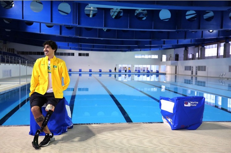 Foto de um ginásio coberto. Há uma piscina olímpica e, à frente, está um rapaz com uma prótese na perna esquerda. Ele veste uma jaqueta dada delegação brasileira, na cor amarela.