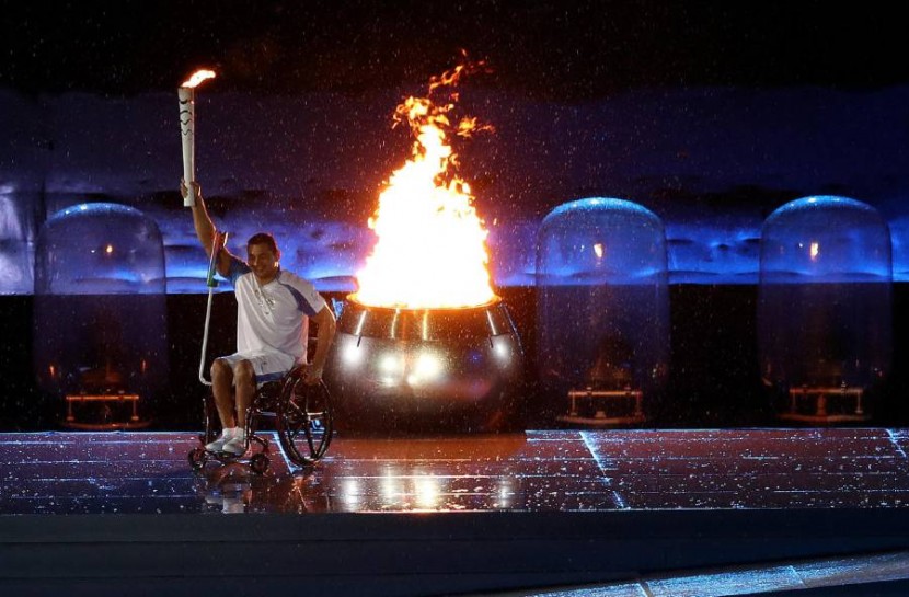Clodoaldo Silva, nadador paralímpico brasileiro, está em sua cadeira de rodas, com e segura a tocha ao lado da pira olímpica acesa. Ele foi o responsável por acender a pira durante a abertura dos jogos.