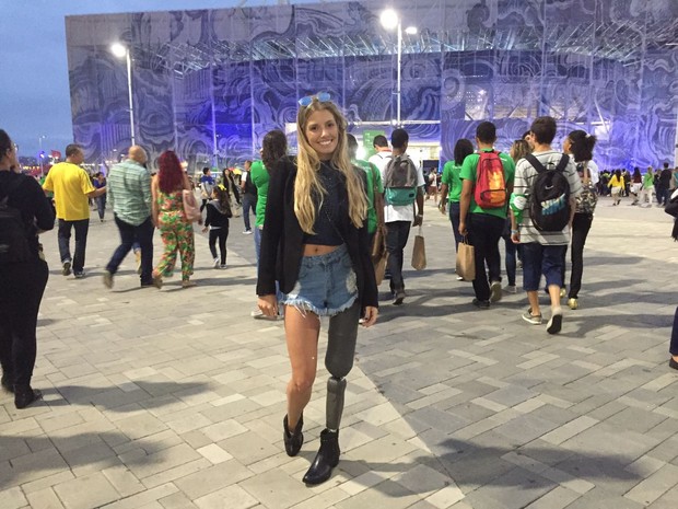 Uma mulher jovem, de longos cabelos loiros, sorri em frente a uma das entradas do Parque Olímpico, com grande movimentação de pessoas ao fundo. Ela usa uma prótese da perna esquerda.