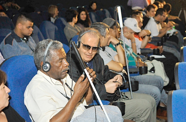 Foto de uma sala de cinema, com várias poltronas. Nelas, estão sentados homens de várias idades, que acompanham a exibição por meio de óculos e fones de ouvido para acessibilidade