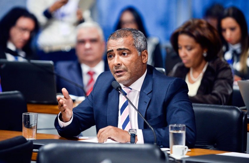 Foto do senado, com diversos políticos sentados ao fundo; o destaque é senador Romário, autor do projeto. Ele usa um terno azul, camisa rosa e uma gravata listrada em azul e rosa e está sentado, falando em um microfone.