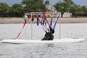 No mar, Mara Gabrilli navega em embarcação adaptada à prática esportiva. Seu corpo está suspenso com auxílio de tiras de segurança atreladas à embarcação.