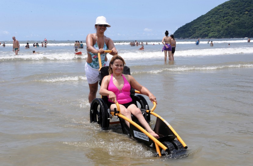 Em uma praia, uma jovem mulher, branca, está sentada em uma cadeira de rodas adaptada à água, com pneus de borracha e suporte para as pernas ficarem esticadas. Uma outra mulher, um pouco mais velha, empurra a cadeira saindo do mar
