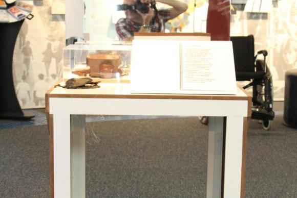 Foto de uma das áreas da exposição, com uma mesa. Nela. há uma encadernação aberta, aparada por um suporte