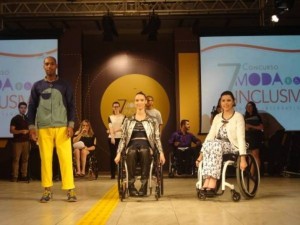 Foto do desfile com roupas inclusivas; três modelos estão em uma passarela. O primeiro modelo é um homem negro e jovem, à esquerda; as duas outras modelos, mulheres brancas e jovens, estão em cadeira de rodas