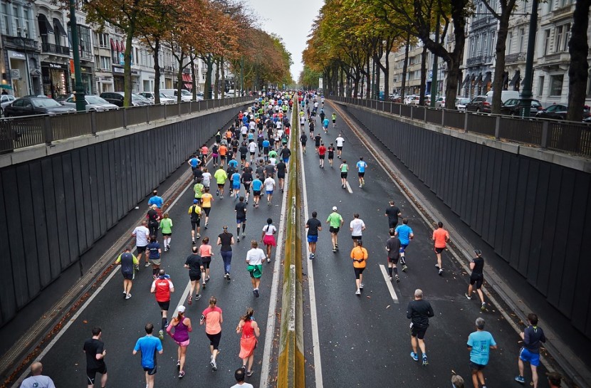 Foto aérea de uma grande avenida asfaltada; pessoas de diversas idades correm por esta avenida, em uma maratona de atletismo