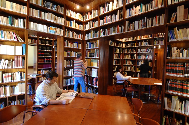 Foto de uma biblioteca, com várias estantes cheias de livros do chão ao teto. Há mesas e cadeiras, com pessoas lendo