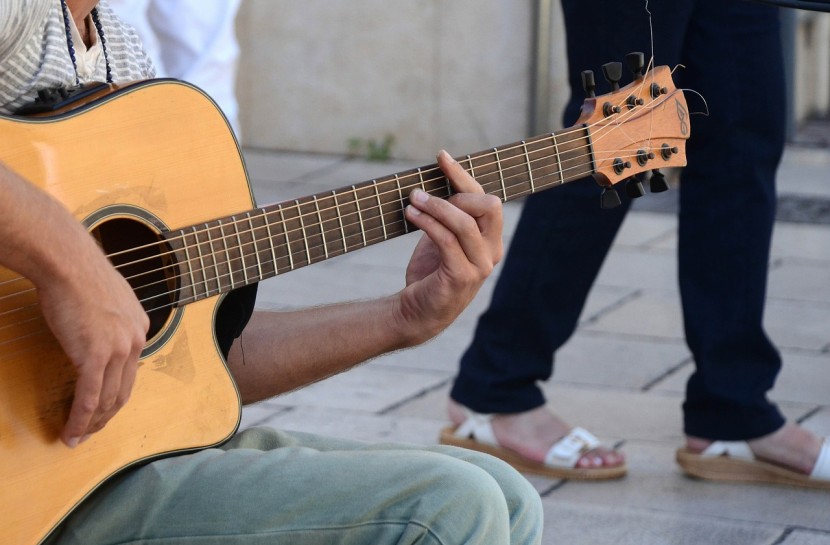 Foto de um homem sentado com um violão no colo. Ele está de perfil, segurando o braço do violão, em primeiro plano.
