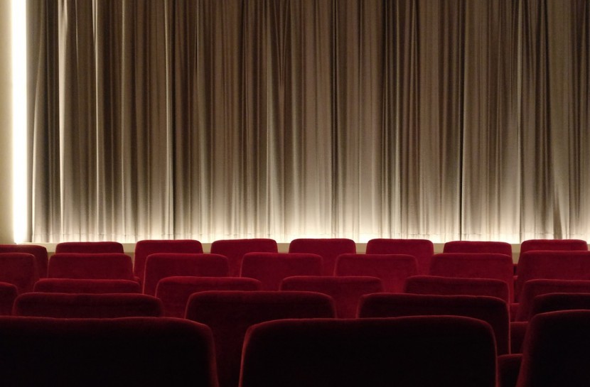 Foto de uma sala de cinema vazia. Ao fundo, uma cortina branca. As cadeiras são vermelhas.