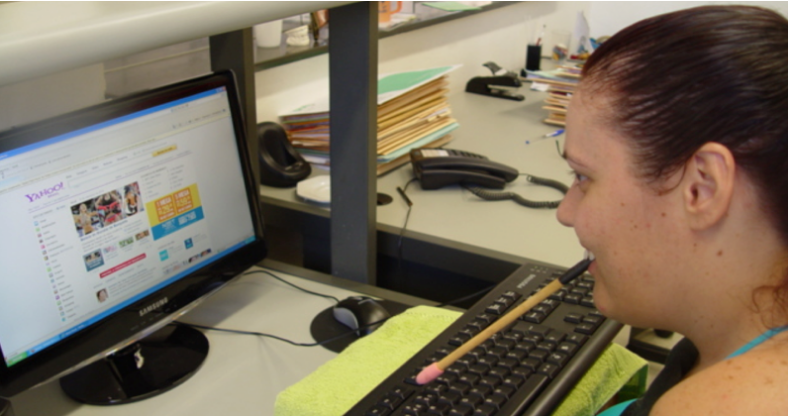 Foto da Joyce, de perfil, usando uma paleta de boca na frente de um computador.