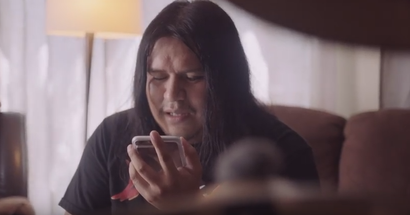 Foto do Carlos, sentado, segurando um telefone celular da Apple próximo à sua boca. Ele é moreno, tem cabelos longos e lisos.