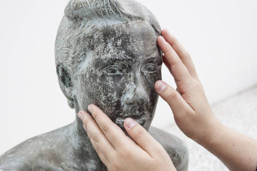 Foto da escultura de um busto, em pedra. Duas mãos a tateiam