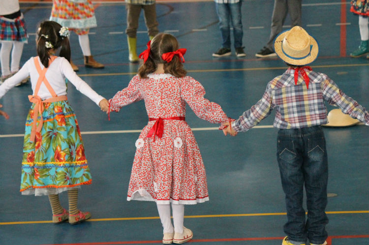 Foto de crianças, duas meninas e um menino, de aproximadamente 8 anos, vestidas com roupas típicas de caipira, de mãos dadas, dançando quadrilha.