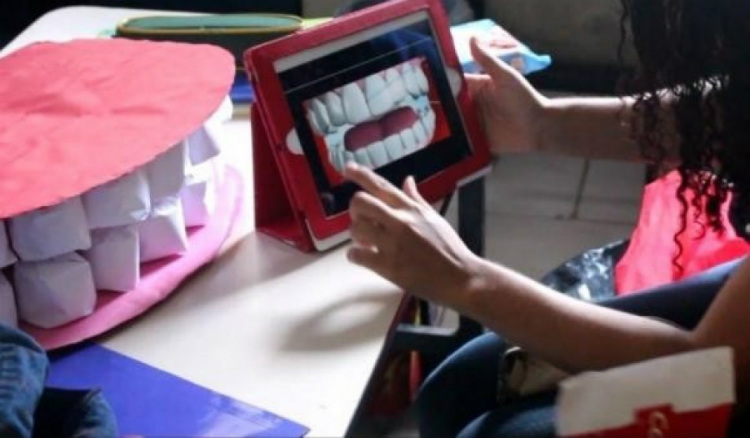 Foto de uma sala de aula. Sobre uma mesa escolar há o protótipo de arcada dentária humana em tamanho grande. À frente, mãos seguram um tablet que mostra esses dentes em uma animação 2D
