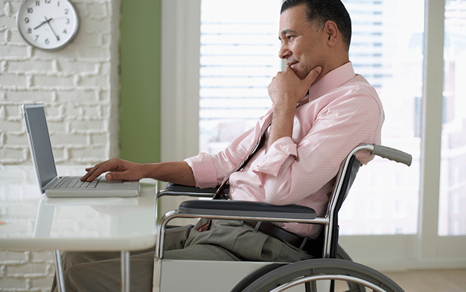 Foto de um senhor negro, sentado em uma cadeira de rodas, olhando curioso a tela de um notebook