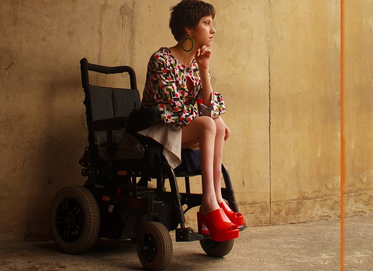 Foto de uma jovem modelo cadeirante posando em uma galpão antigo, com um vestido. Ela olha na direção direita com semblante contemplativo