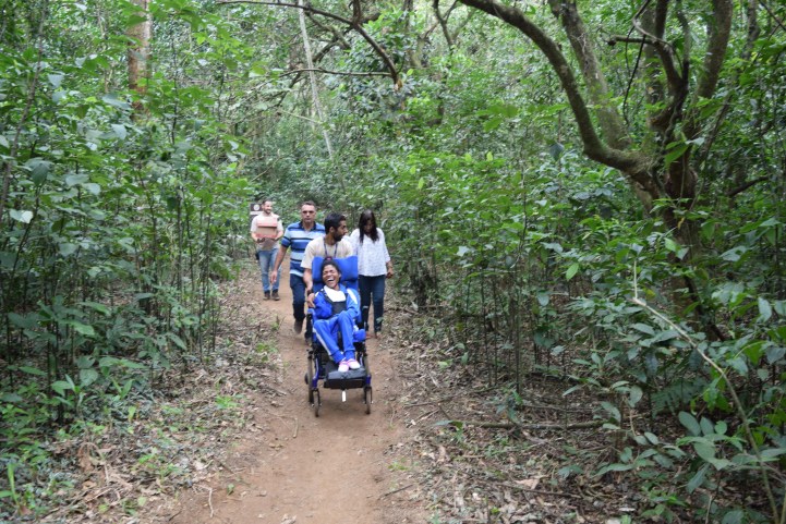 Foto de uma trilha repleta de árvores, com um caminho em terra ao meio. Cinco pessoas, de diferentes idades, estão andando. À frente, está uma jovem negra, cadeirante, que sorri.