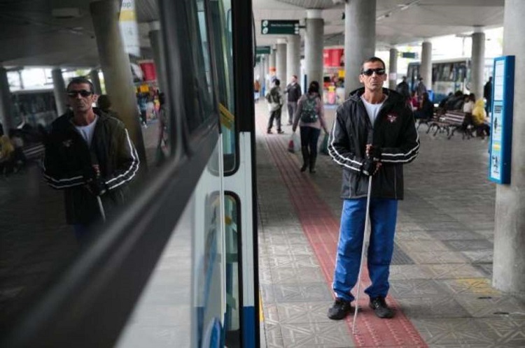 Foto de um terminal rodoviário. Um homem de meia idade, cego, com óculos escuros e uma bengala. está de pé, do lado de um ônibus