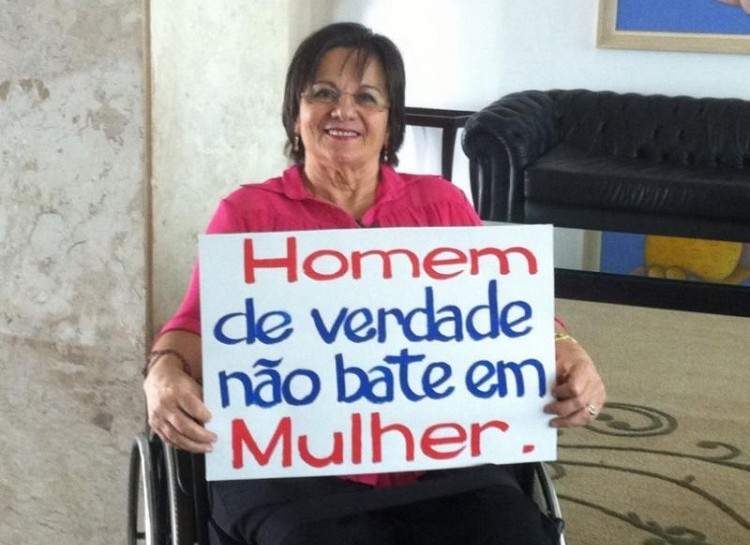 Foto de Maria da Penha, sentada em uma cadeira de rodas, segurando um cartaz com os dizeres: Homem de verdade não bate em mulher