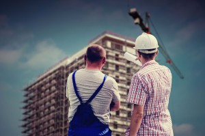 Foto de dois homens em pé olhando um prédio. Um deles está usando macacão e outro está usando blusa social. Um deles está com capacete branco na cabeça.