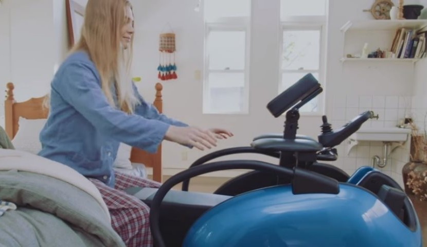 Foto de uma jovem mulher loira de pijamas, sentada na beira da cama. Perto dela, há uma cadeira motorizada, com uma base azul que indica o assento