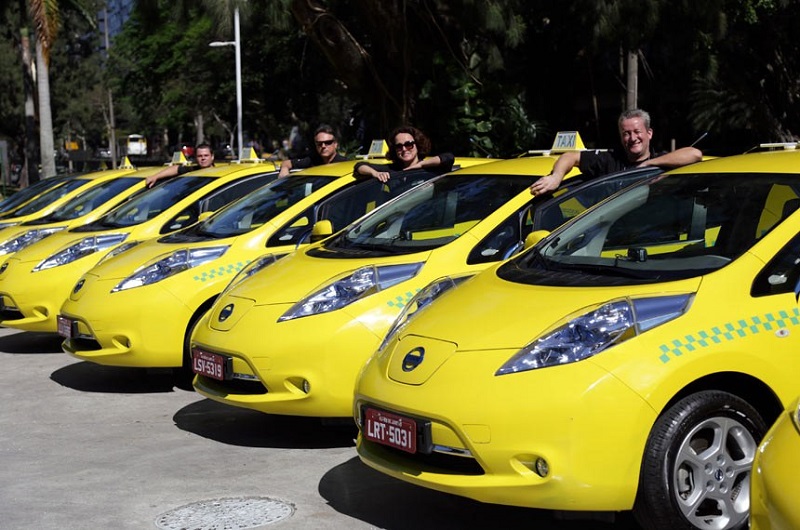 Foto de uma frota de táxis amarelos, em fila, com pessoas de diversas idades, ao lado das porta do motorista de cada veículo