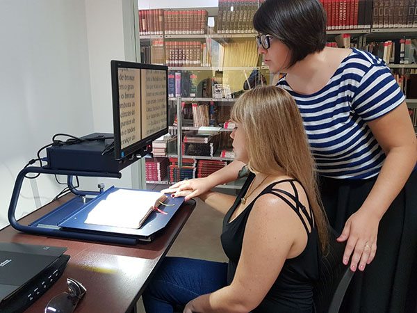 Foto de duas mulheres em uma biblioteca, com dispositivos acessíveis para leitura à frente.