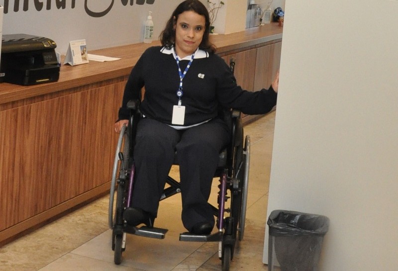 Foto da entrada do Inclui CIEE, com uma jovem cadeirante na porta. Ela está uniformizada, usa um crachá e sorri
