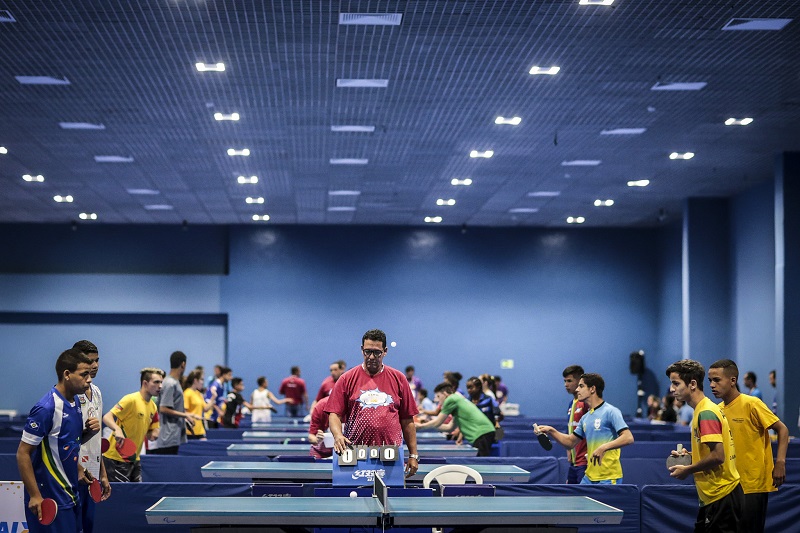 Foto de um grande salão com diversas mesas de tênis de mesa. Em destaque, um instrutor orienta dois garotos de aproximadamente 14 anos, que seguram raquetes