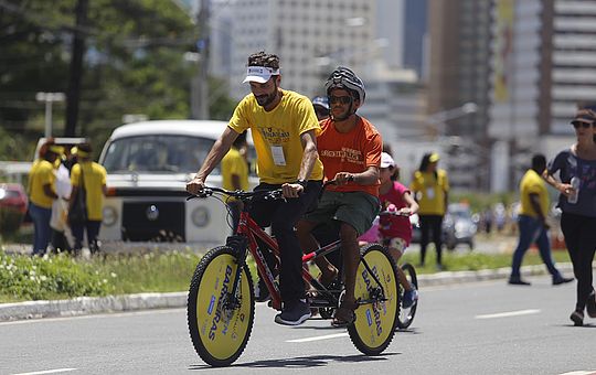 Foto de uma avenida, com dois homens jovens de capacete guiando uma bicicleta adaptada, com dois bancos e dois guidões