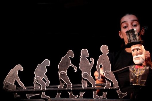 Foto em close de marionetes articulados em fila, indicando a evolução do macaco até o homem. No lado direito, há um boneco de Charles Darwin, um senhor com cartola e longa barba branca