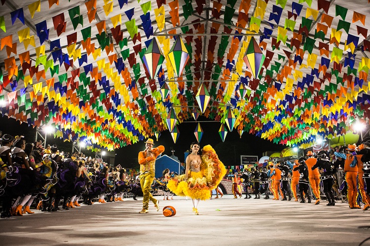 Foto em plano aberto da Festa de São João de Caruaru. Entre bandeirinhas, há um casal com trajes de frevo dançando em um grande corredor, rodeado de pessoas