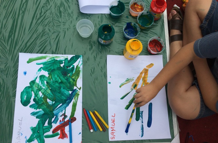 Foto de duas mãos de crianças sujas de tintas coloridas, segurando um pincel e pintando sobre folhas em branco em uma mesa