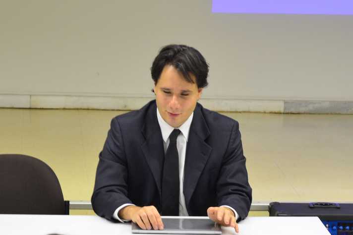 Foto de Leonardo Coscarelli, um homem jovem, branco, com cabelos escuros na altura do queixo. Ele usa terno e gravata azuis e camisa branca, está sentado atrás de uma mesa