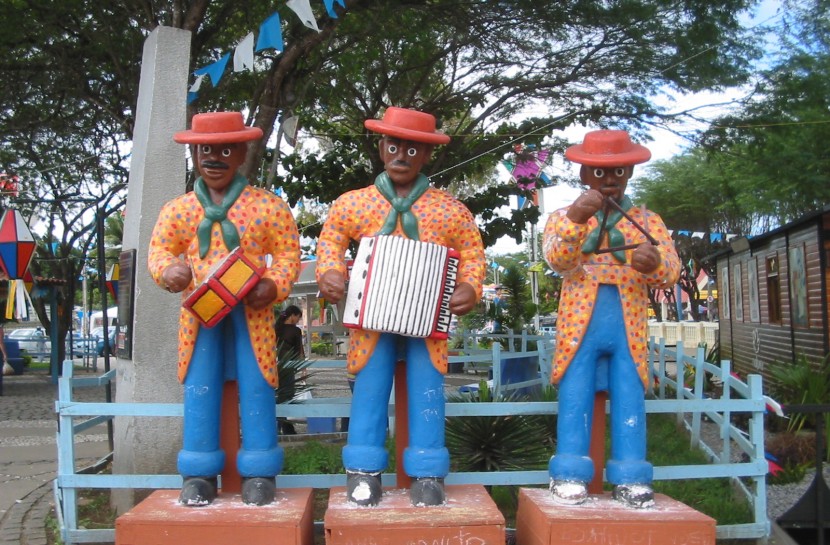 Foto de uma escultura de cerâmica de um Típico trio de forró pé-de-serra: com três homens tocando zabumba, sanfona e triângulo. Eles usam chapéu vermelho, balzer laranja e calça azul e estão em uma praça de Caruaru