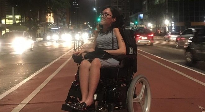 Mulher jovem em cadeira de rodas na Avenida Paulista. Ela é morena, tem cabelos na altura dos ombros e usa um vestido listrado