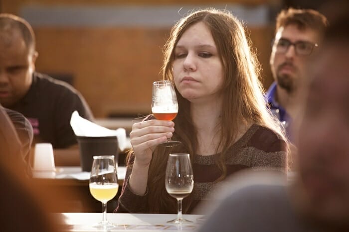 Jovem mulher segurando uma taça de cerveja próxima ao rosto. Ela é branca e tem cabelos na cor castanho claro