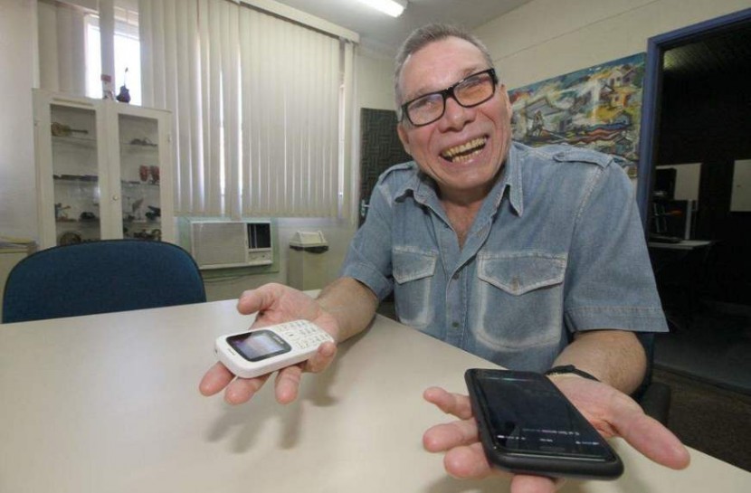 Foto de Gilson, um senhor de aparentemente 60 anos, com óculos e segurando um celular e um smartphone