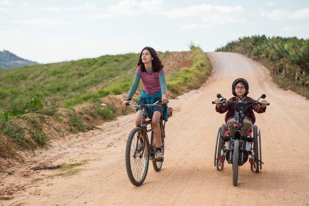 Cena do filme Sobre Rodas. Um garota e um garoto andam de bicicleta em uma estrada de terra. A bicicleta do garoto é adaptada