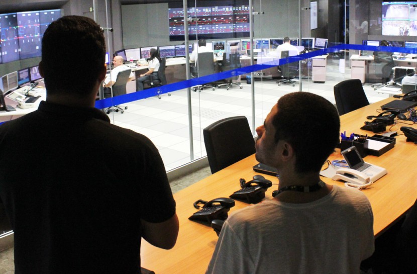 Foto de Lucas e um funcionário do metrô no Centro de Controle Operacional do Metrô, de costas, olhando através de uma parede de vidro para uma sala repleta de painéis eletrônicos e computadores